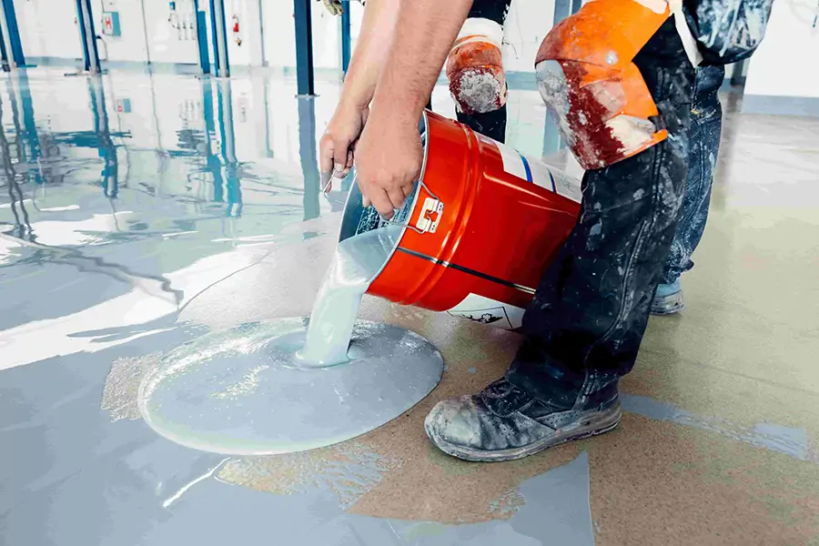 Worker pouring gray epoxy floor coating onto a commercial concrete floor.